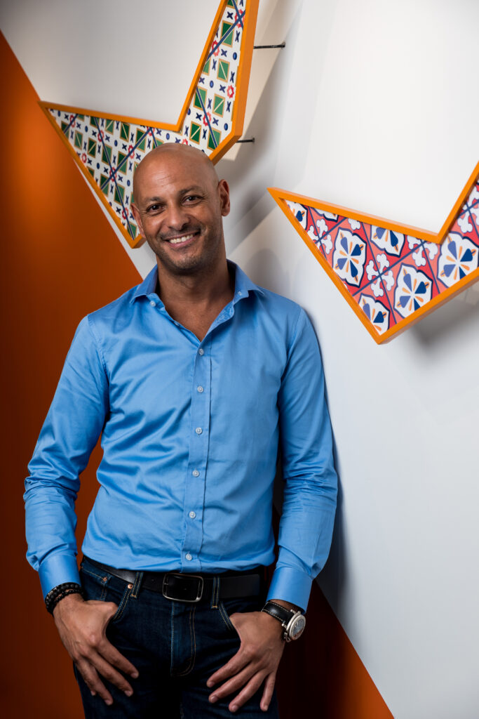 A man in a light blue shirt and dark jeans stands smiling against a white and orange wall decorated with colorful, geometric mosaic patterns.