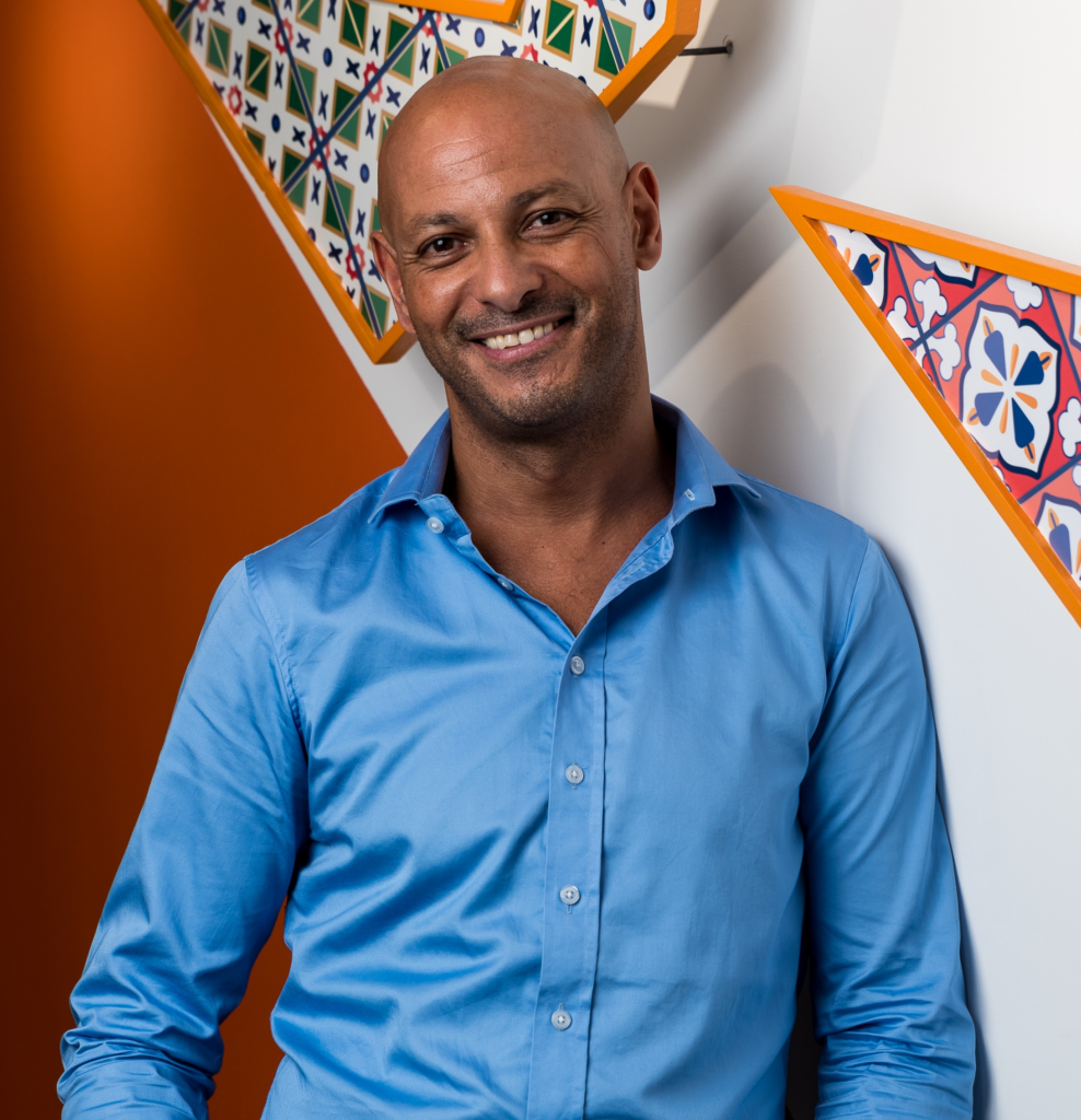 A smiling man in a light blue shirt stands against a wall with colorful, patterned geometric decorations and an orange accent.