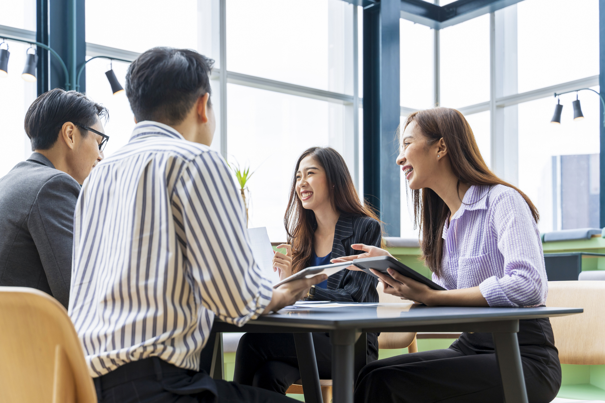 Four young adults sit around a table in a bright office, smiling and talking while holding tablets and papers, suggesting a collaborative meeting or discussion.