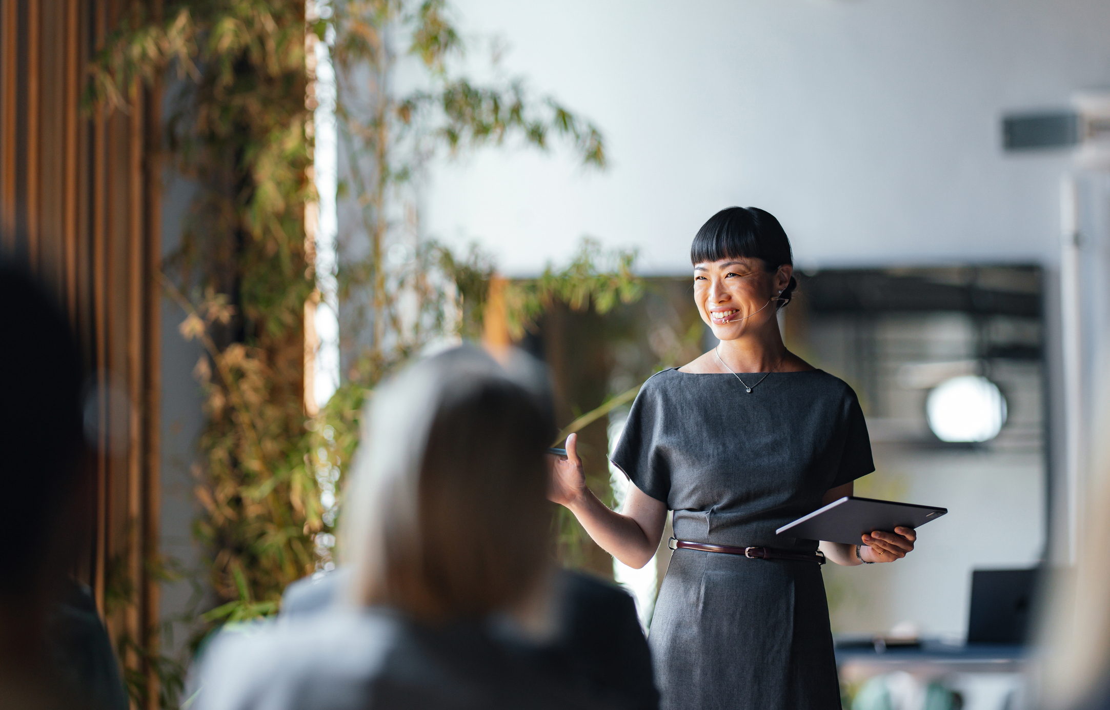 A woman in a gray dress smiles and gestures while holding a tablet, giving a presentation to a seated audience in a modern, plant-filled room.