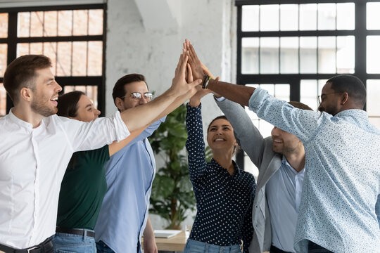 A group of six diverse colleagues stand in a circle indoors, smiling and giving each other a high-five, celebrating teamwork and success in a bright office setting.