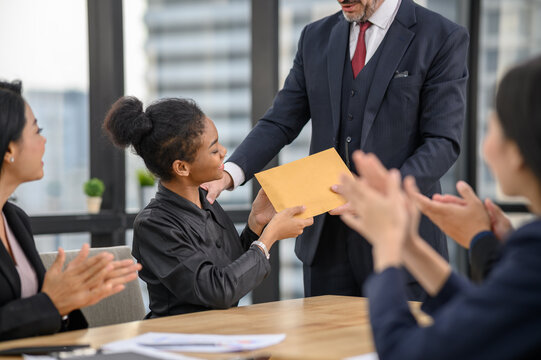 A woman receiving a brown envelope from a man in a suit while seated at a table, as others sit nearby clapping in a modern office setting.
