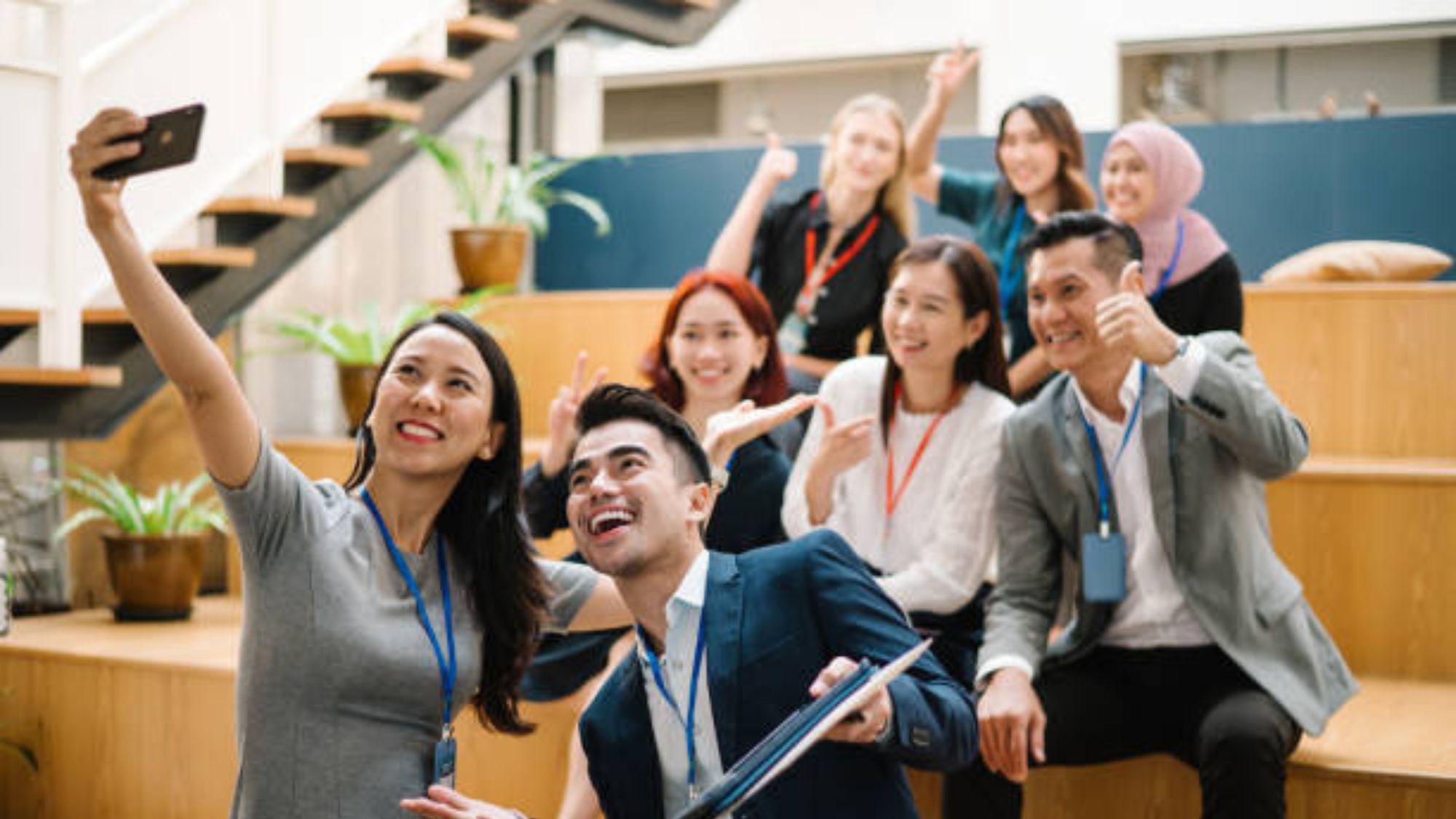 A group of seven coworkers smile and pose for a selfie together on wooden steps in a modern office, all wearing name badges and displaying cheerful expressions.