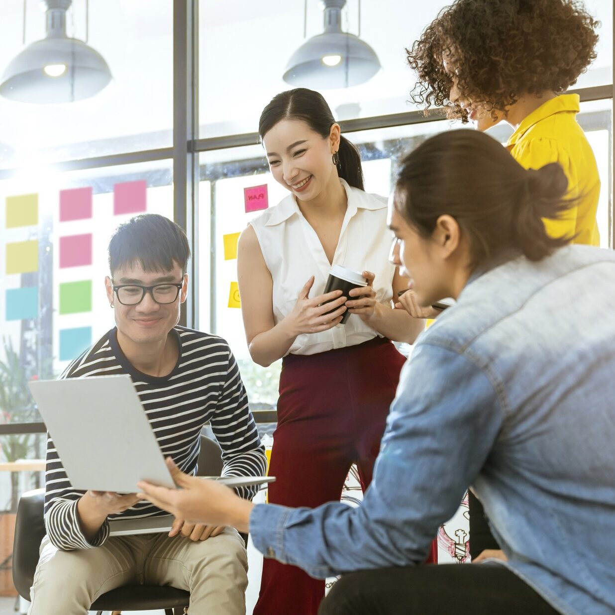 Four people gather in a modern office, smiling and looking at a laptop. Colorful sticky notes cover the glass wall behind them, and one person holds a notebook while another gestures to the laptop screen.
