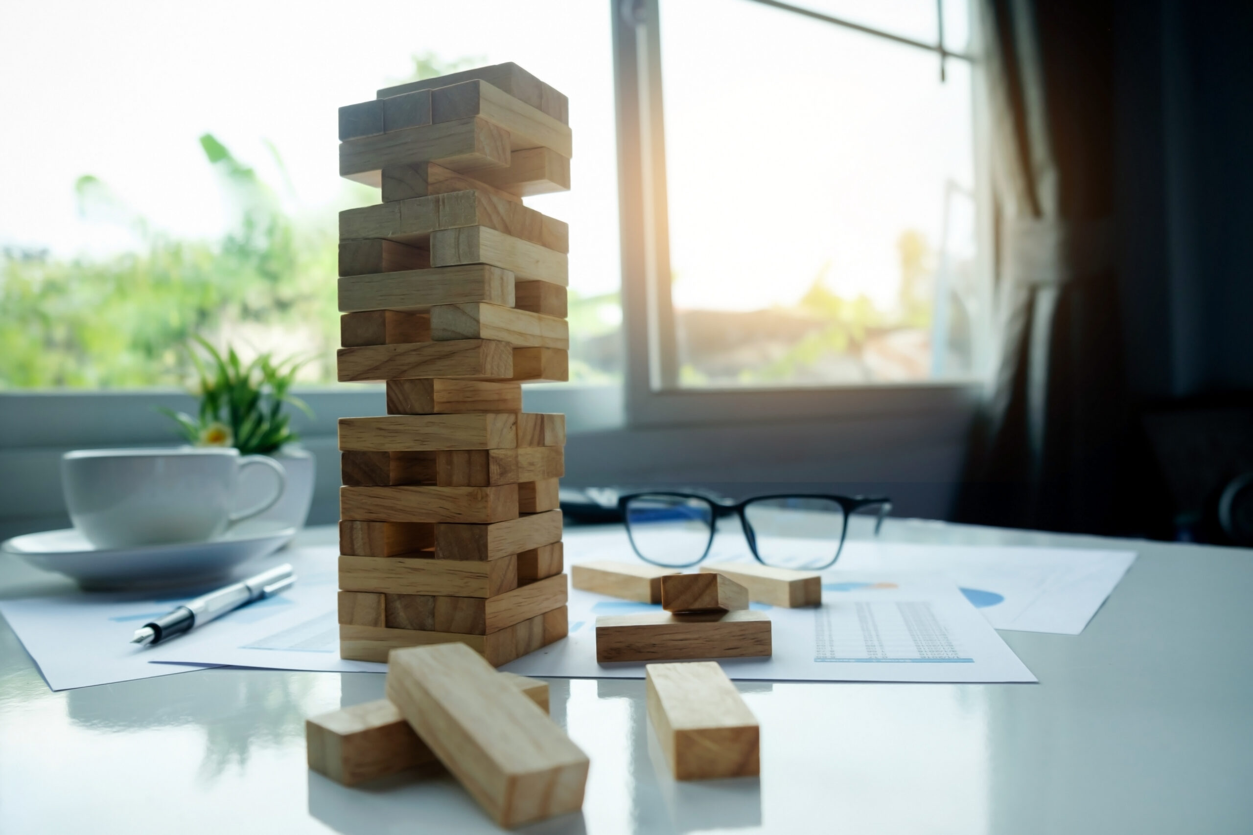 A Jenga tower stands on a white table, surrounded by fallen wooden blocks. Nearby are eyeglasses, a cup and saucer, a pen, and papers. Sunlight streams through a window, and a small plant is visible in the background.