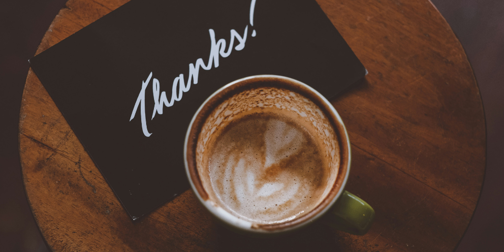 A green cup with frothy coffee sits on a wooden table beside a black card inscribed with Thanks! in elegant white script, serving as a token of recognition and appreciation.