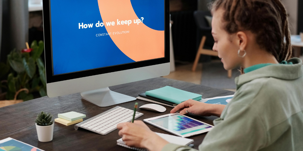 A person sits at a desk, a computer screen displaying How do we keep up? Constant evolution while they hold a stylus, examining color swatches on a tablet. Nearby, office supplies and a small plant create an inspiring workspace that fuels creativity for designing sales incentive games.