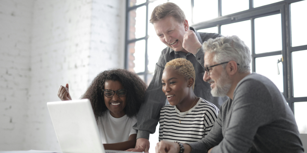A group of four people, two women and two men of different ages, are gathered around a laptop in a bright room with large windows. They are smiling and appear to be motivated as they collaborate on boosting sales strategies displayed on the screen.