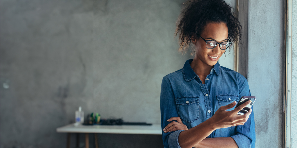 A person wearing glasses and a denim shirt stands near a window, smiling while looking at their smartphone. The background is softly blurred, showing part of a desk with items on it against a neutral wall.