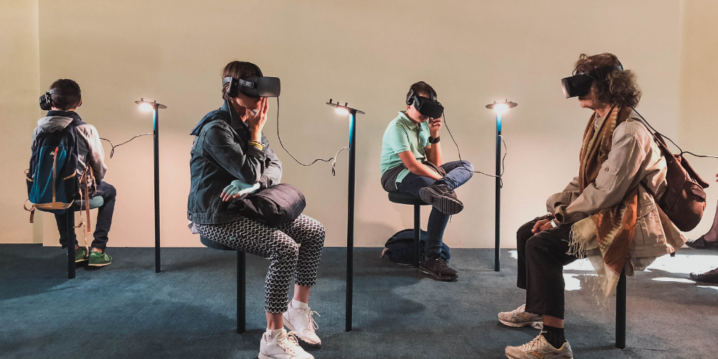 Four people sit on stools wearing VR headsets and headphones in a dimly lit room. Each person is connected to a stand with wires. The backdrop is a plain, light-colored wall.