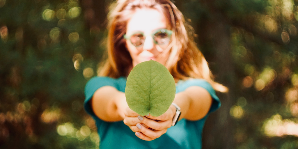A person with long hair holds a large green leaf towards the camera, wearing sunglasses and a blue shirt. The background is a lush and blurred forest, creating a vibrant and natural atmosphere.