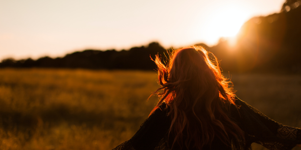 A person with long red hair stands in a sunlit field, arms outstretched. The golden light of the setting sun casts a warm glow over the landscape, creating a serene and peaceful atmosphere.