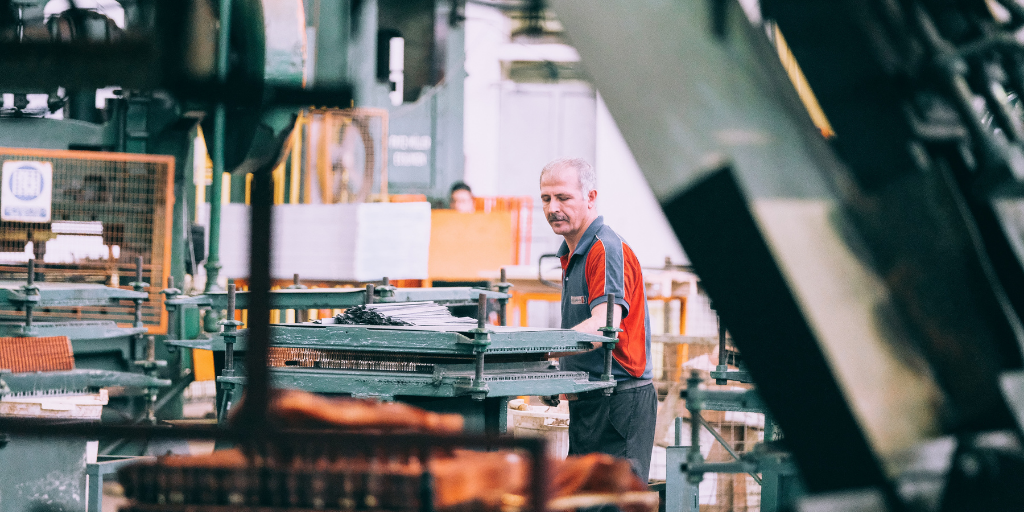 A man in a factory setting, wearing a red and black shirt, operates machinery surrounded by metal structures and components. The environment appears industrial, with various equipment and materials in the background.