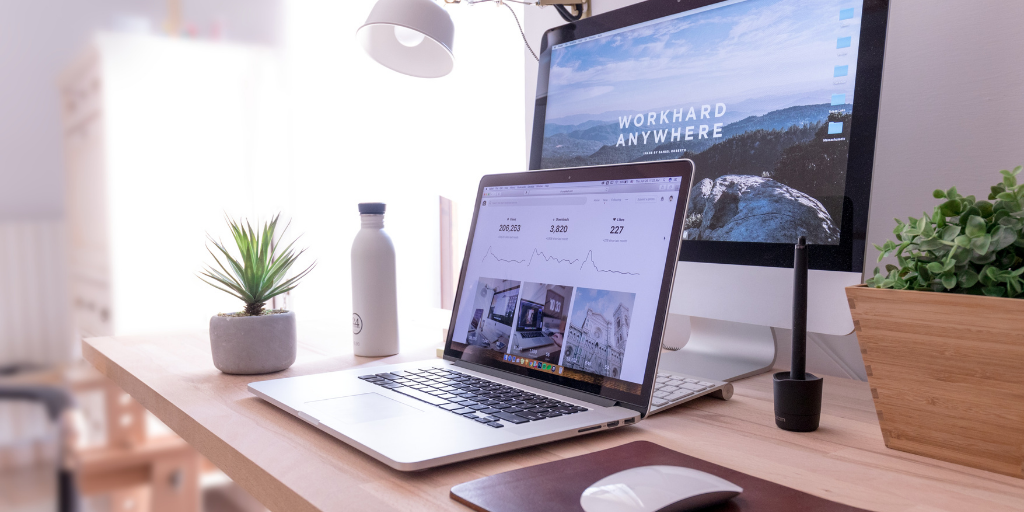 Modern workspace with a laptop displaying analytics, an iMac with Work Hard Anywhere on the screen, a small plant, water bottle, pen tablet, and a mouse on a wooden desk, illuminated by a desk lamp.