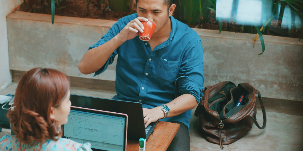 A person in a blue shirt drinks from a can while working on a laptop, prioritizing their social health through casual collaboration. Another employee sits across with an open laptop, and a brown bag rests on the floor. They are surrounded by plants in this relaxed indoor setting.