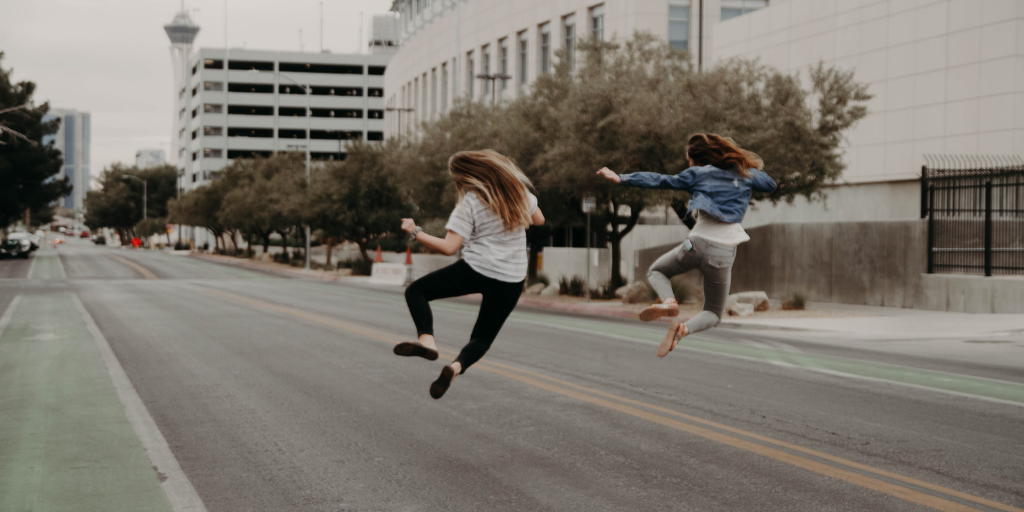 Two people jump joyfully in the middle of a deserted street. They wear casual clothes, with trees and modern buildings lining the road. The atmosphere is carefree and lively.