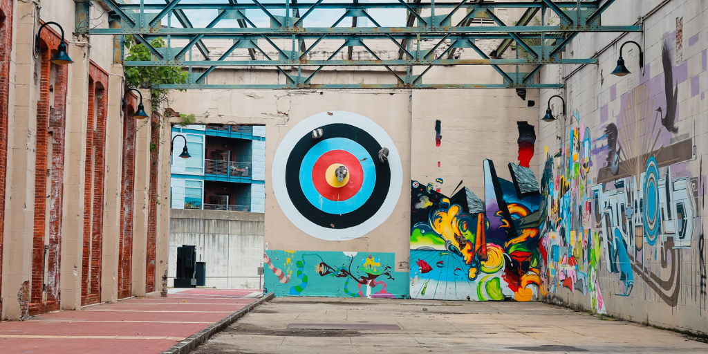 Outdoor industrial space with vibrant street art on walls. A large, colorful target design is central, with abstract, colorful graffiti to the right. Exposed beams and industrial lights overhead, modern building partially visible in the background.