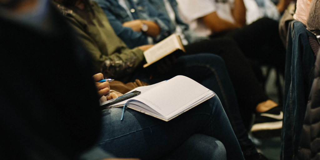 People seated in a row, focusing on an event. Some hold open books or notepads on their laps. One person holds a pen over a notebook. The scene suggests a lecture or seminar setting. The atmosphere is attentive and contemplative.