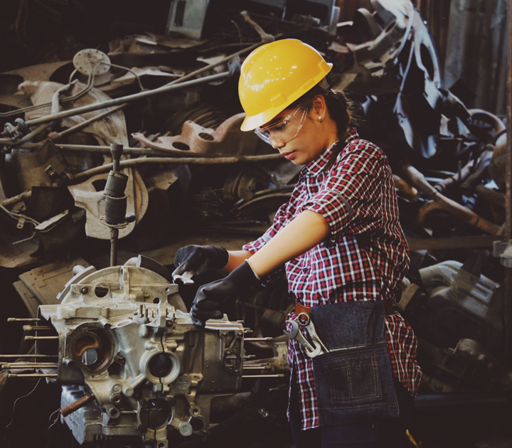 Person in a plaid shirt and yellow hard hat works on machinery in an industrial setting. They wear safety goggles and gloves, with various tools attached to their apron. Background features metal parts and equipment.