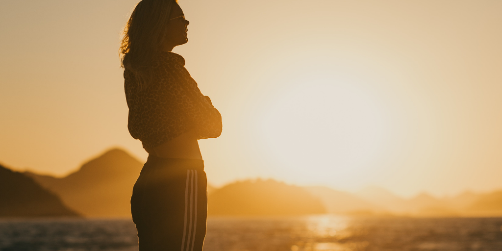 Silhouette of a person standing near water during a vibrant sunset. Their posture is contemplative, and mountains are visible in the background. The scene is warm and serene, with the sun low on the horizon.