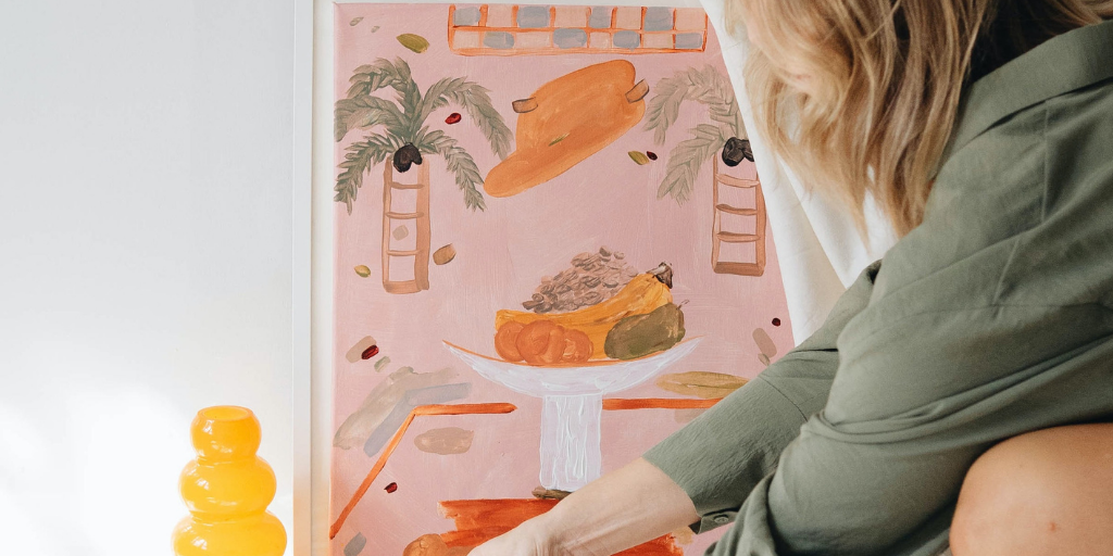 A person in a green shirt is creatively adjusting a pink-framed artwork of palm trees and a fruit-filled bowl. Nearby, an innovative yellow glass object adds a touch of flair to the surface.