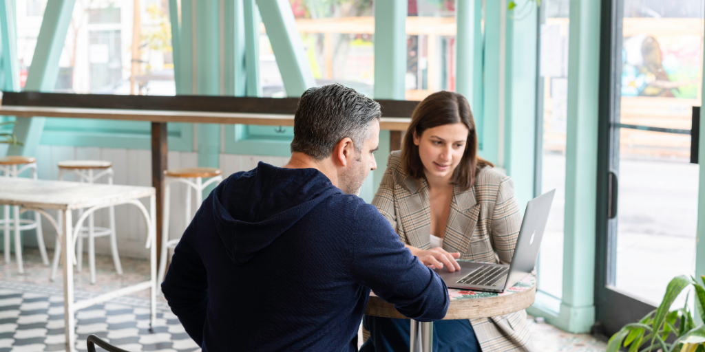 Two people sitting at a round table in a café with teal walls. They are focused on a laptop in front of them. The man wears a navy sweater, and the woman wears a plaid blazer. High stools are visible in the background.