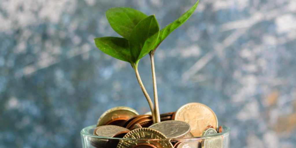 A small plant with green leaves grows from a pile of assorted coins in a glass jar, set against a blurred, textured blue-gray background.