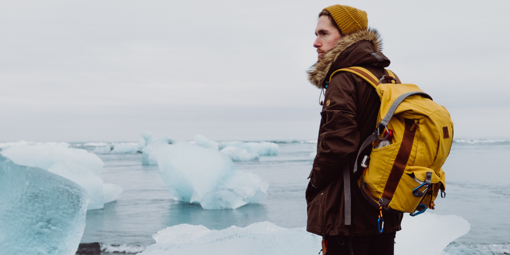 A person stands on a snowy shore, facing the side, wearing a brown coat with fur trim, a yellow beanie, and a yellow backpack. Large ice formations float in the water under an overcast sky.