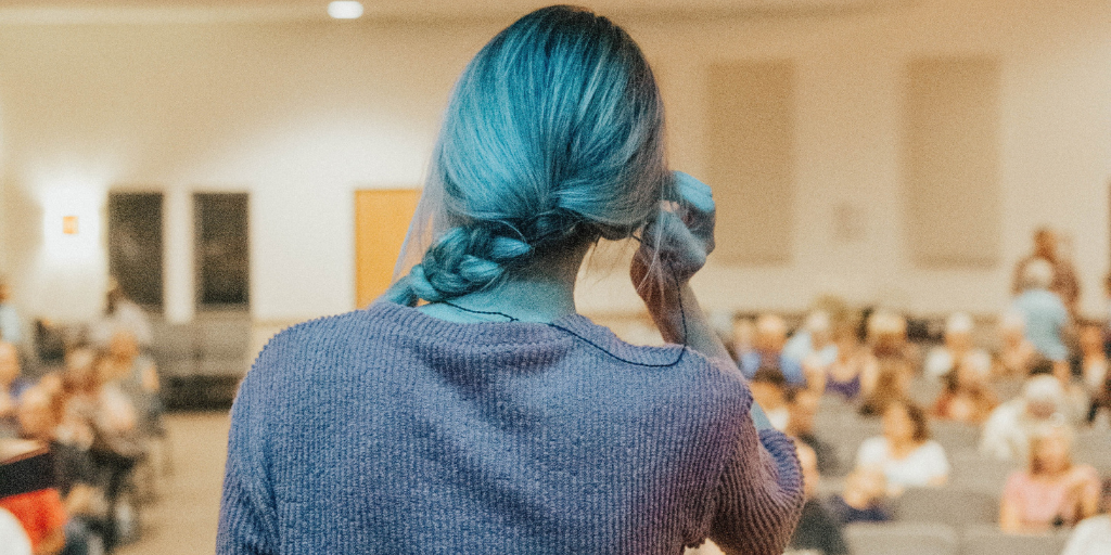 A person with blue hair tied in a braid stands on stage, facing an audience in a dimly lit room. The audience is seated, and the focus is on the speaker from behind. The atmosphere suggests a presentation or lecture.
