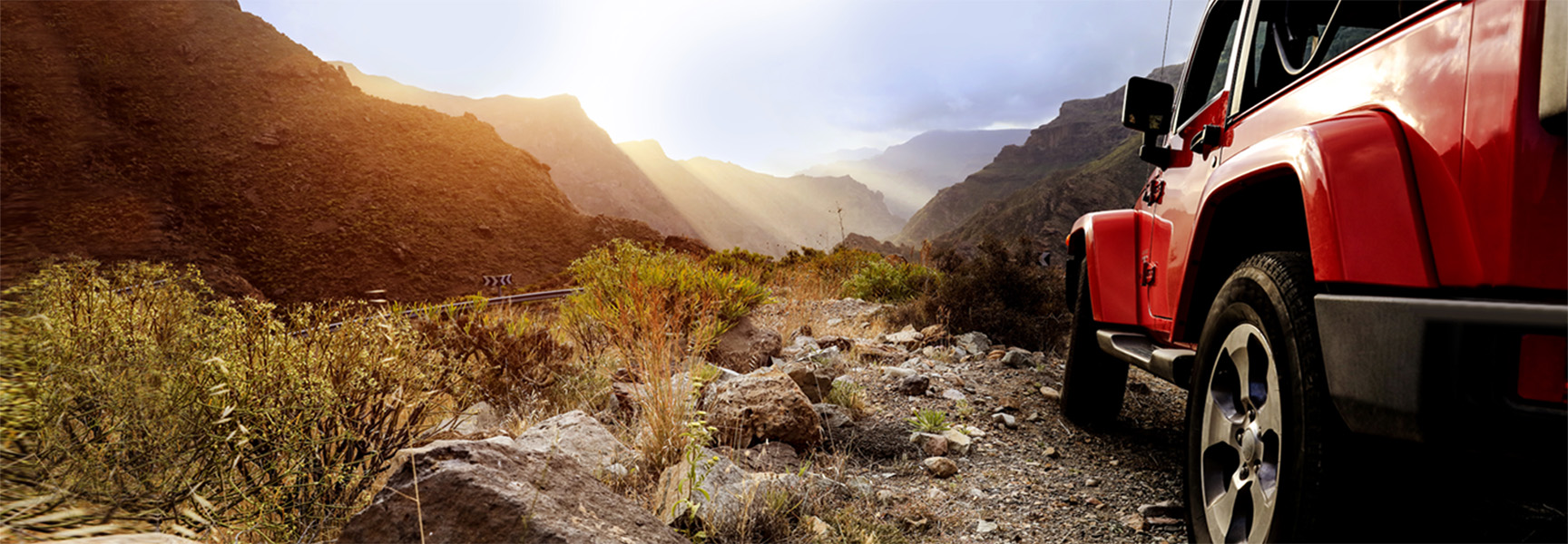 A red off-road vehicle navigates a rugged, rocky path through mountains at sunset, with rays of sunlight streaming through clouds and illuminating the landscape. Vegetation is sparse along the trail.