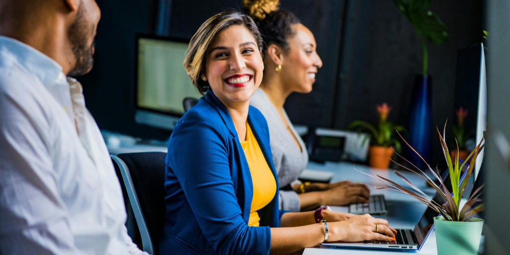 A woman in a blue blazer and yellow top smiles brightly while sitting at a desk in an office. She is in front of a laptop, discussing channel incentive strategies. Two colleagues, a man and another woman, are seated beside her, focused on their computers. A plant adds a touch of greenery to the desk.