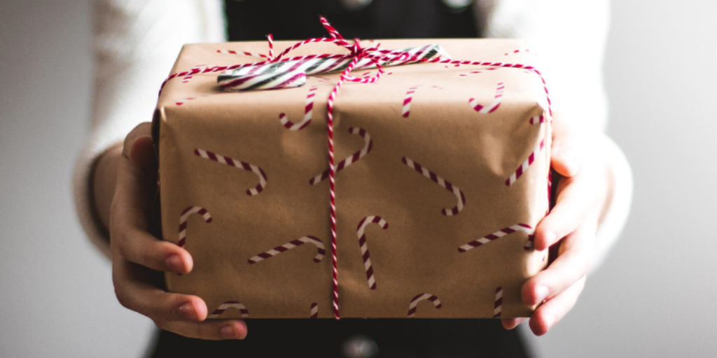A person holding a gift wrapped in brown paper with red and white candy cane patterns, tied with a red and white string bow. The background is blurred.