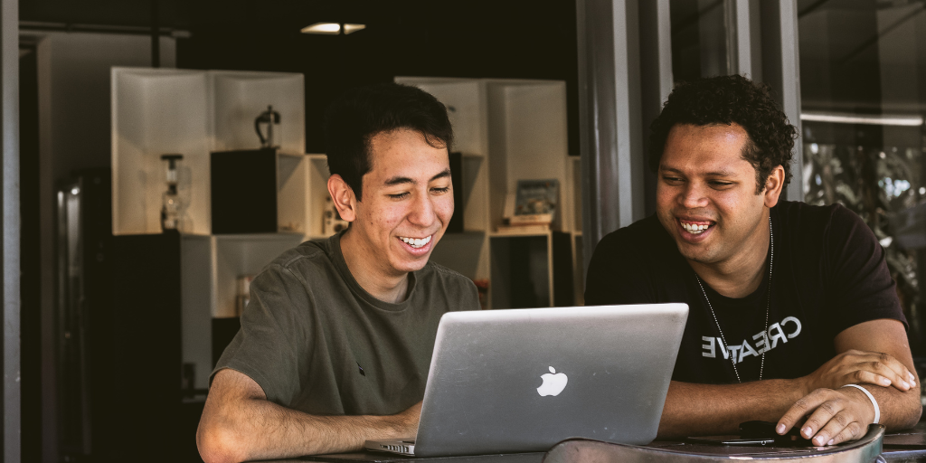 Two people sitting at an outdoor table are smiling while looking at a laptop. Engaged and focused on the screen, they seem to be enjoying their work, perhaps reviewing employee performance data. A modern interior adds a touch of style in the background.