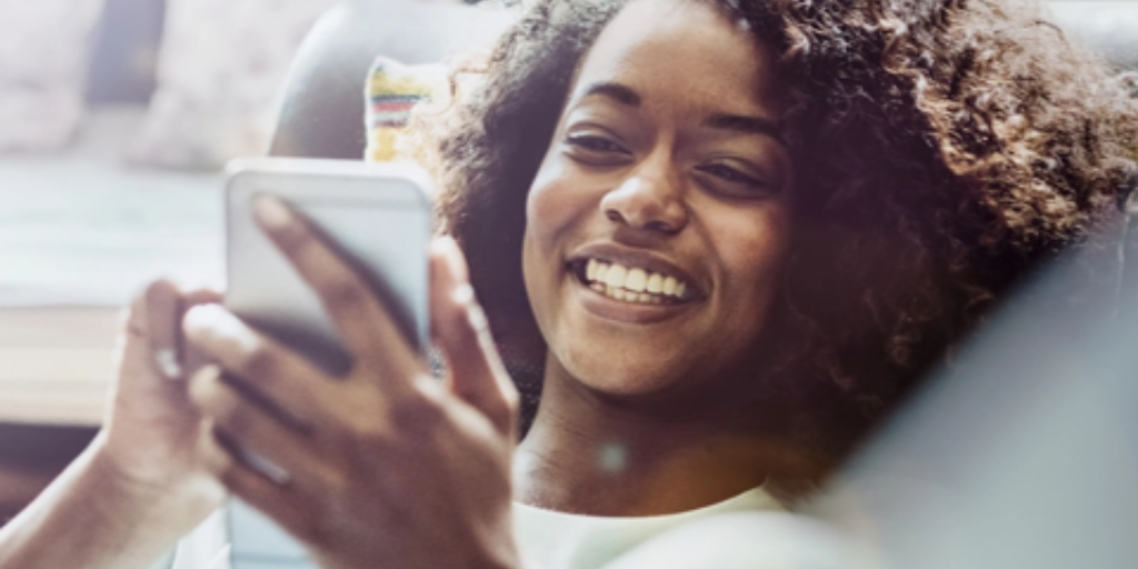 A person with curly hair is smiling while checking event communications on a smartphone. Seated indoors in a casual white shirt, they appear relaxed as sunlight softly illuminates the scene.
