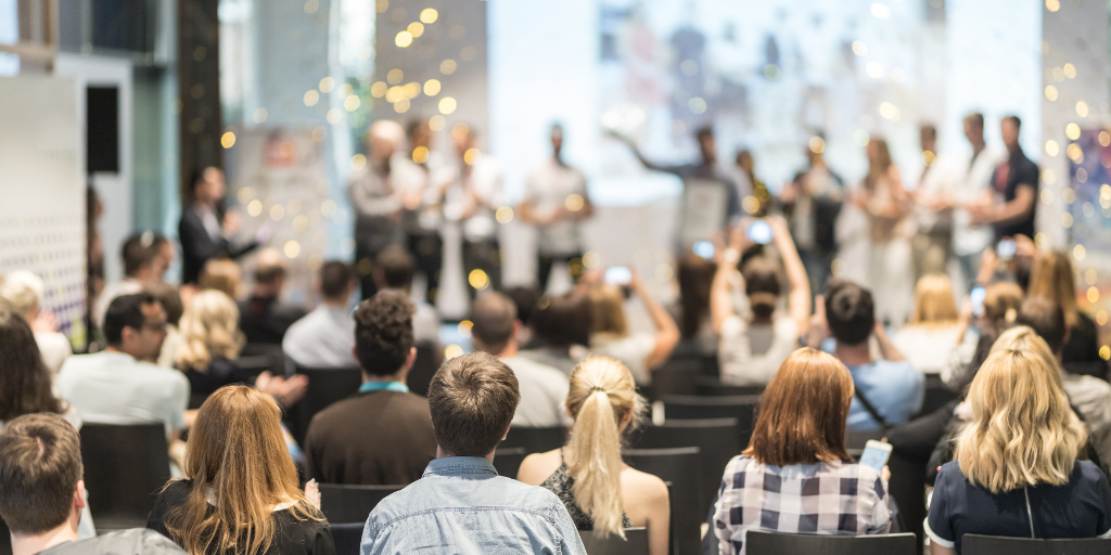 A large group of people seated in a conference room, facing a blurred stage. Some, perhaps from the employee service team, are taking photos or videos with their phones. The room is decorated with white and gold lights, creating a festive atmosphere.