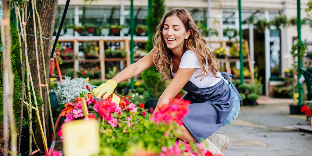 A woman wearing a blue apron and gardening gloves is tending to colorful flowers at an outdoor nursery. She is smiling and surrounded by various plants in pots and planters, with a greenhouse structure visible in the background.