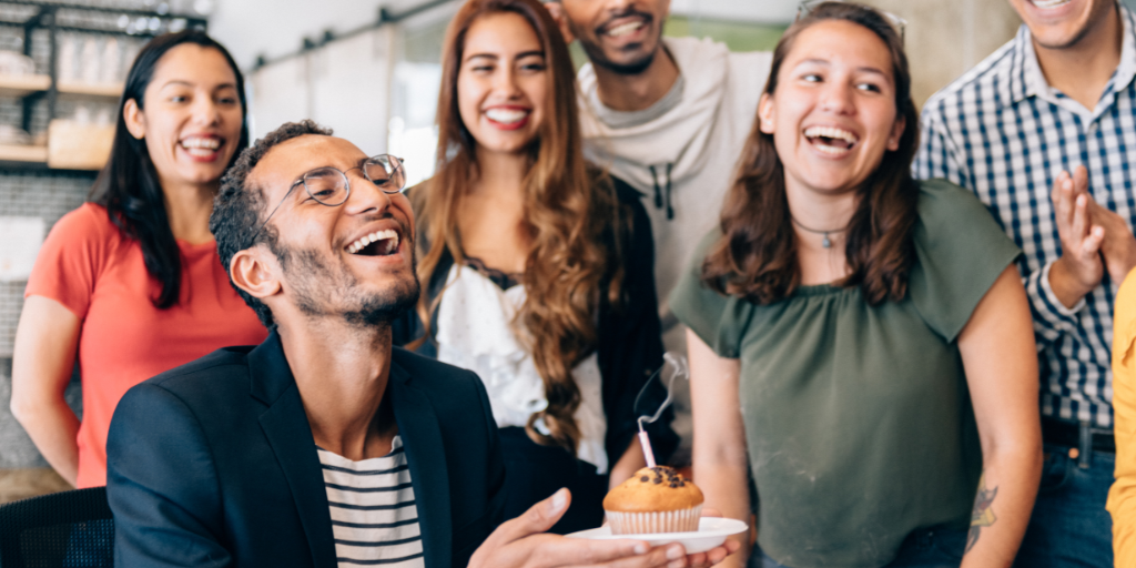 A group of people gathered together, smiling and laughing in celebration. One person in the foreground holds a small cake with a lit candle, marking employee milestones. The atmosphere is joyful and festive.