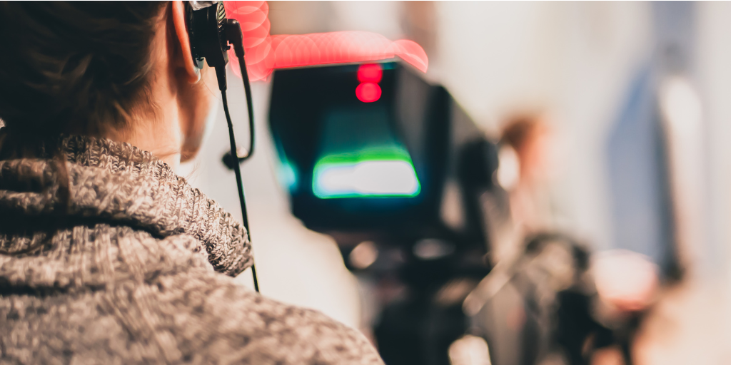 Person in a gray sweater operates a professional video camera on a set. The camera is slightly out of focus, highlighting the person’s concentration on the task. The background is blurred, emphasizing the action of filming.
