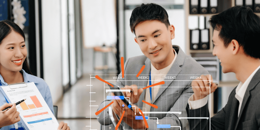 Three people are in an office discussing a chart displayed on a transparent board. One is holding a document with graphs, another points at the chart, and the third smiles, engaging in the conversation. They appear to be planning or analyzing data.