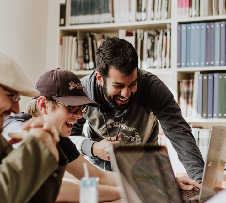 Three people sit and laugh around a table with laptops in a library or office, embodying great work culture. They seem to share something funny on the screen, while shelves filled with books and binders stand in the background.