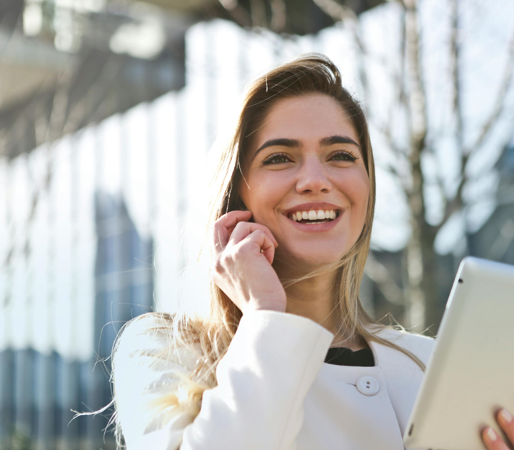 A woman is smiling and looking up while engaging in a lively conversation on her smartphone, appreciation quotes subtly reflected in her expression. She holds a tablet and stands outdoors, clad in a light-colored jacket, with a glass building and trees framing the scene.