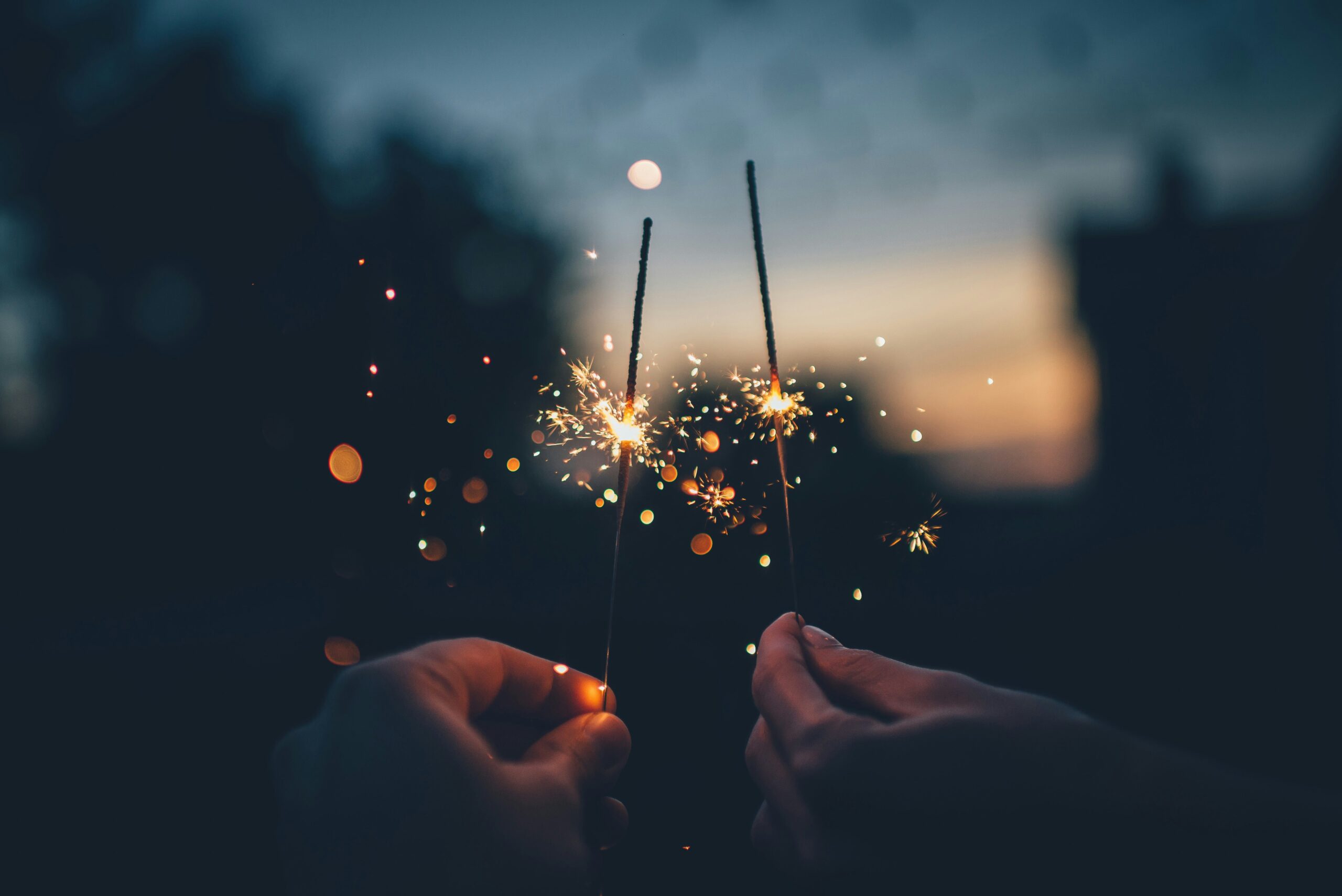 Two hands holding lit sparklers against a blurred, dusk background create a festive atmosphere reminiscent of measuring corporate events. The bright, glowing sparks emit a warm, soft glow with a bokeh effect from distant lights.