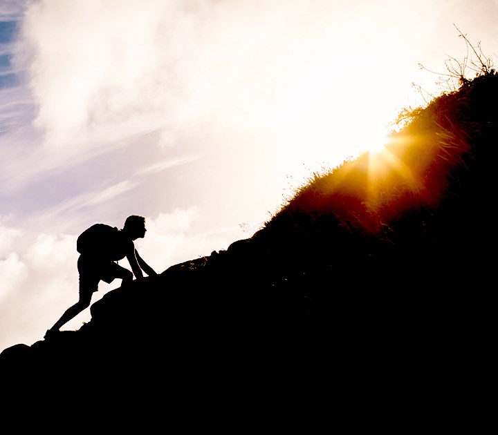 Silhouette of a person hiking uphill with a backpack against a bright sunrise or sunset.