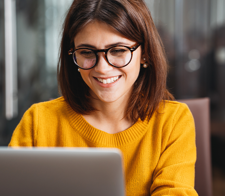 A woman with shoulder-length brown hair and glasses is smiling at her laptop, embodying employee motivation. She wears a yellow sweater and sits in an indoor setting with a blurred background, exuding positivity and focus.