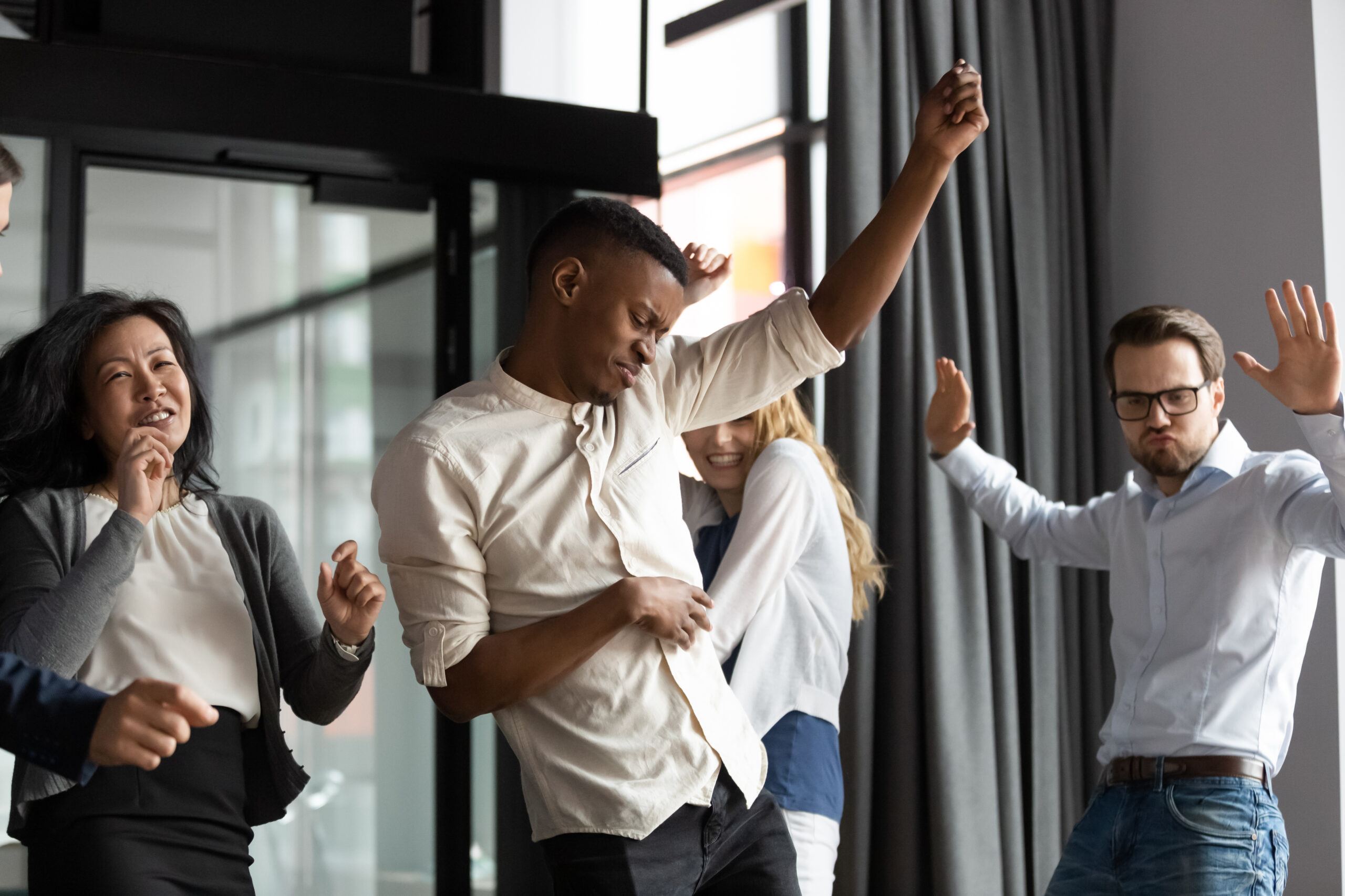 A group of five people dances energetically in an office setting, embodying employee motivation. They appear joyful, expressing themselves through lively movements. Natural light filters through large windows in the background, and curtains are visible.