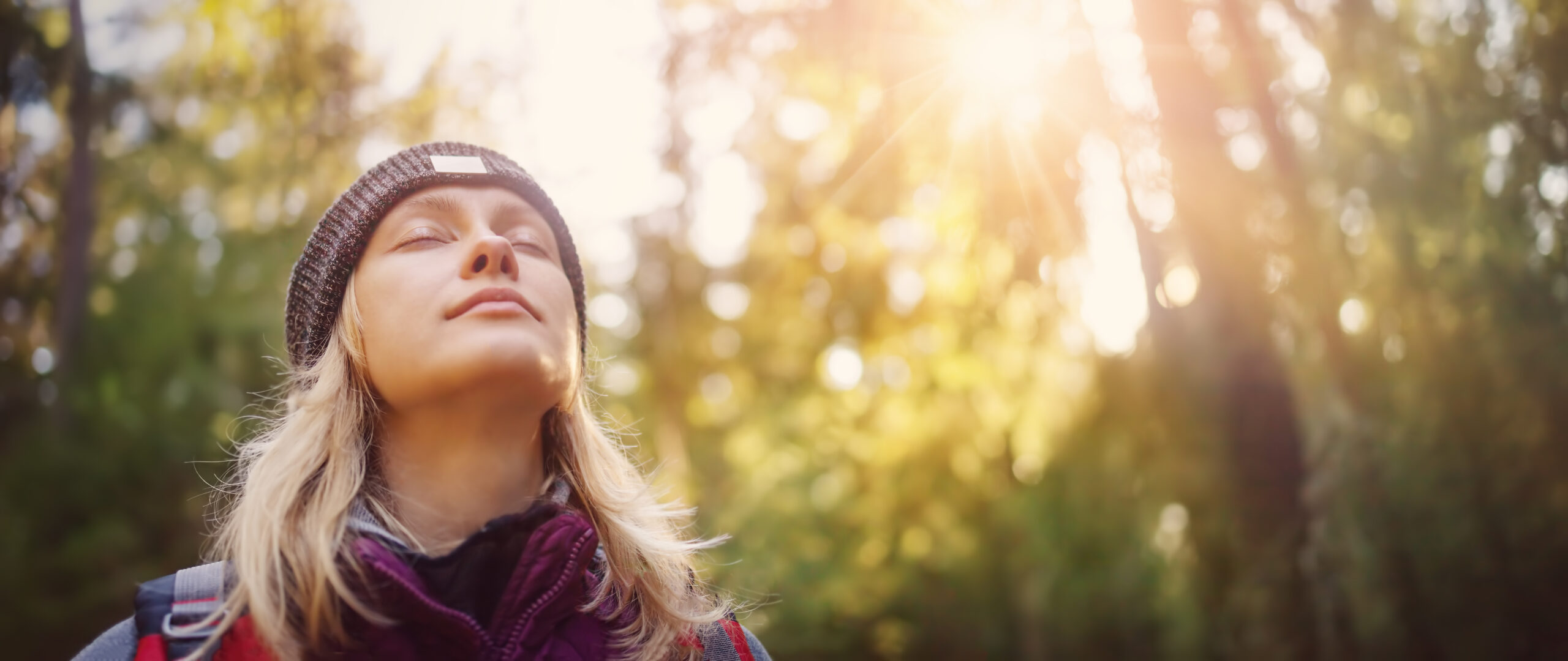 Woman with closed eyes and a content expression stands in a sunlit forest. She wears a knit hat and warm clothing. Sunlight filters through the trees, creating a serene and peaceful atmosphere.