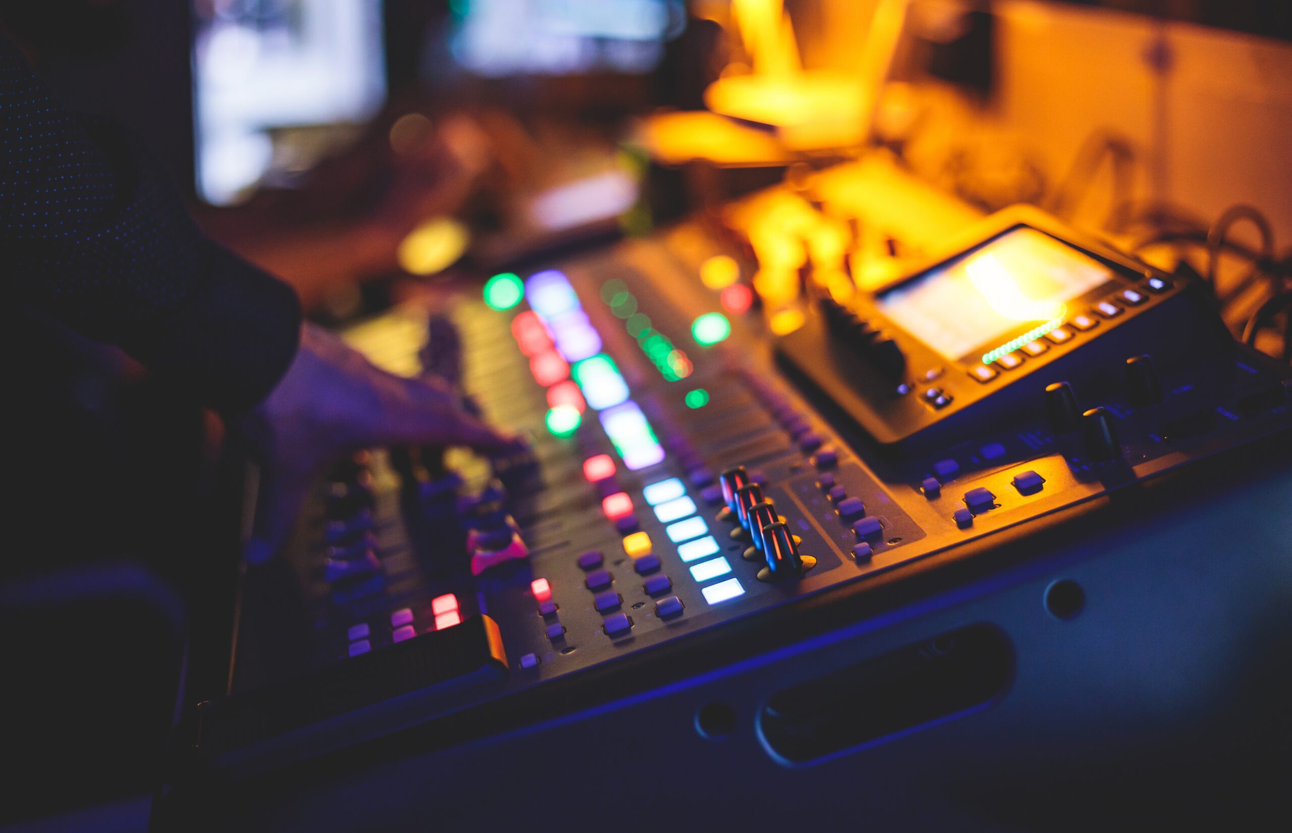 A persons hand adjusting controls on a colorful, illuminated sound mixing board in a dimly lit room. Various buttons and knobs are lit in different colors, with a small display screen visible on the right.