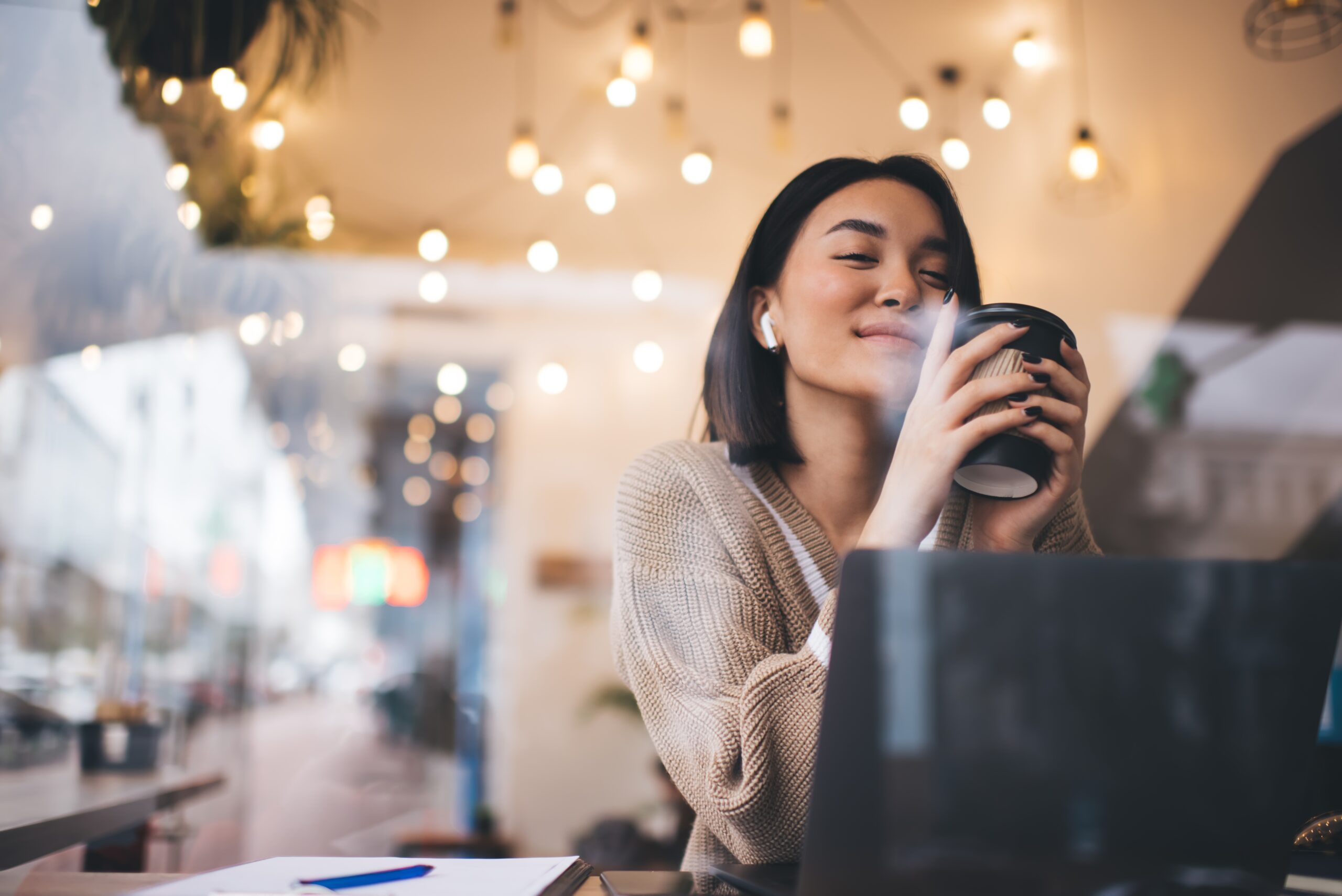 A woman with shoulder-length hair is sitting in a cozy cafe, holding a to-go cup and smiling contently. Shes near a laptop, with warm, hanging lights in the background, creating a relaxed atmosphere.