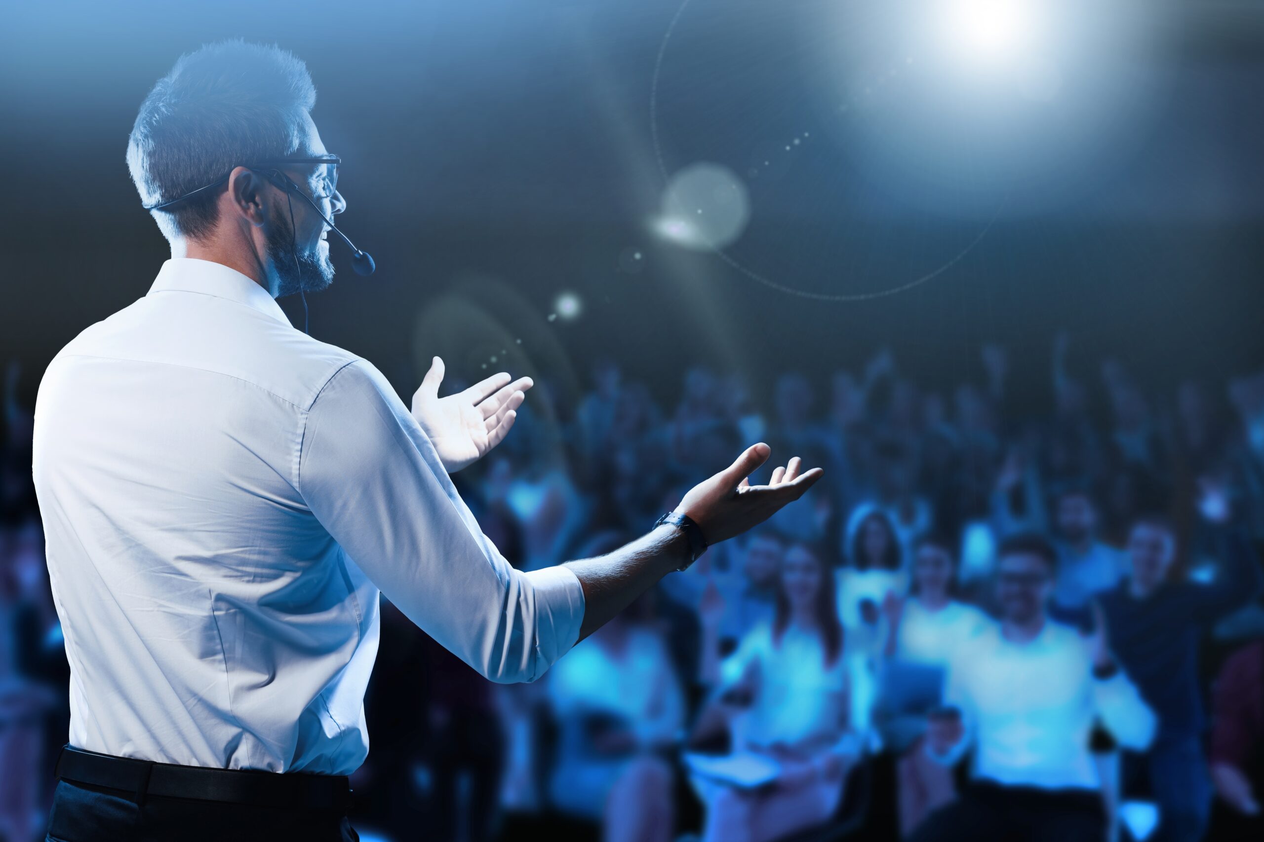 A man in a white shirt, wearing glasses and a headset, gestures while speaking to an audience in a dimly lit auditorium. The focus is on him, with the audience blurred in the background. Bright stage lighting highlights the scene.