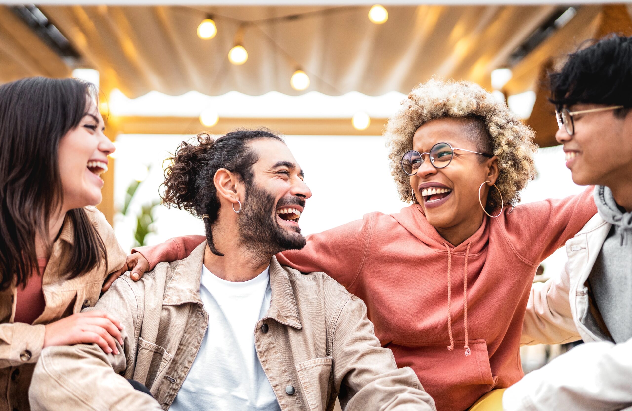 A group of four diverse friends, participating in a recognition programme, sits closely together, laughing joyfully. They wear casual clothes and are under ambient string lights, creating a warm, friendly atmosphere.